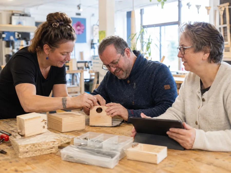 Begeleider en cliënten werken samen in de houtwerkplaats, met het cliëntdossier op een tablet